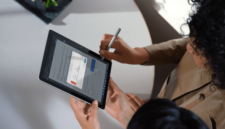 A bird's eye view shows two people, one holding the Surface Go 3 in tablet mode and the other signing a document on it with the Surface Pen 