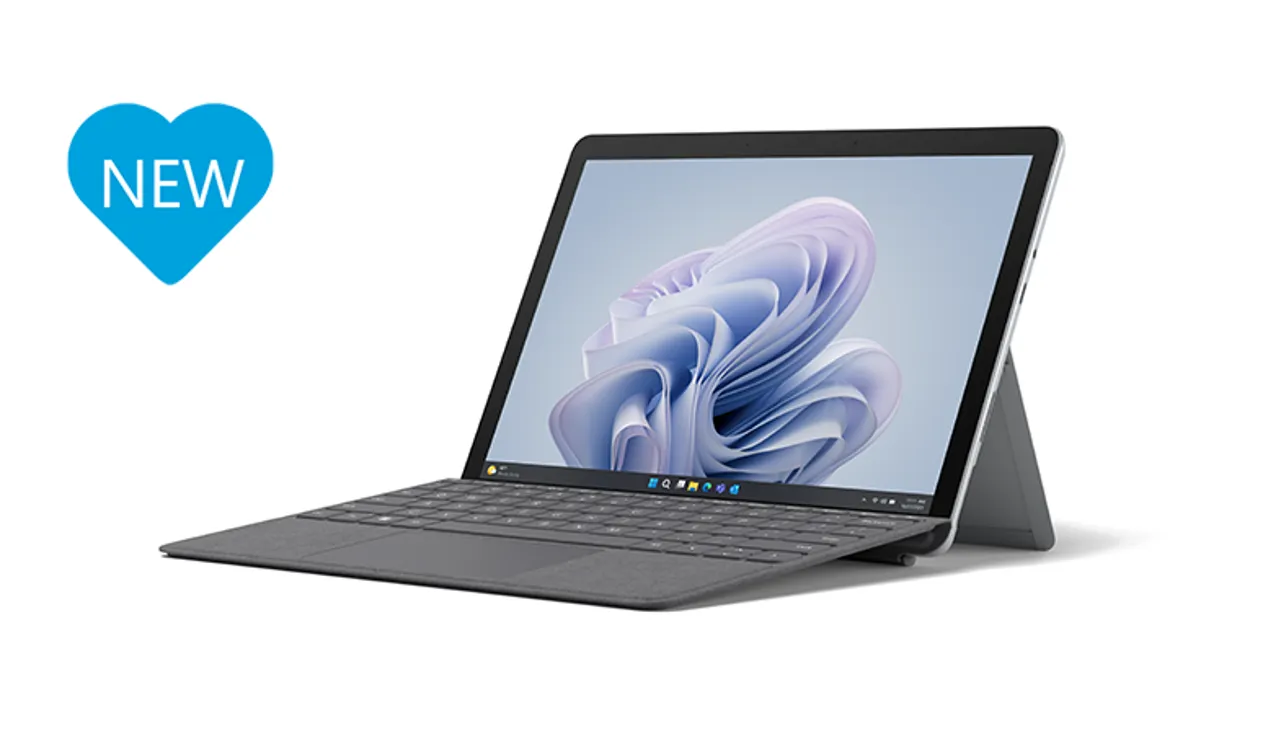 Das Surface Go 4 in Platin inklusive Type Cover in Schwarz  im Laptop-Modus aus seitlicher Perspektive mit blauem Herz links neben dem Surface auf dem NEU steht