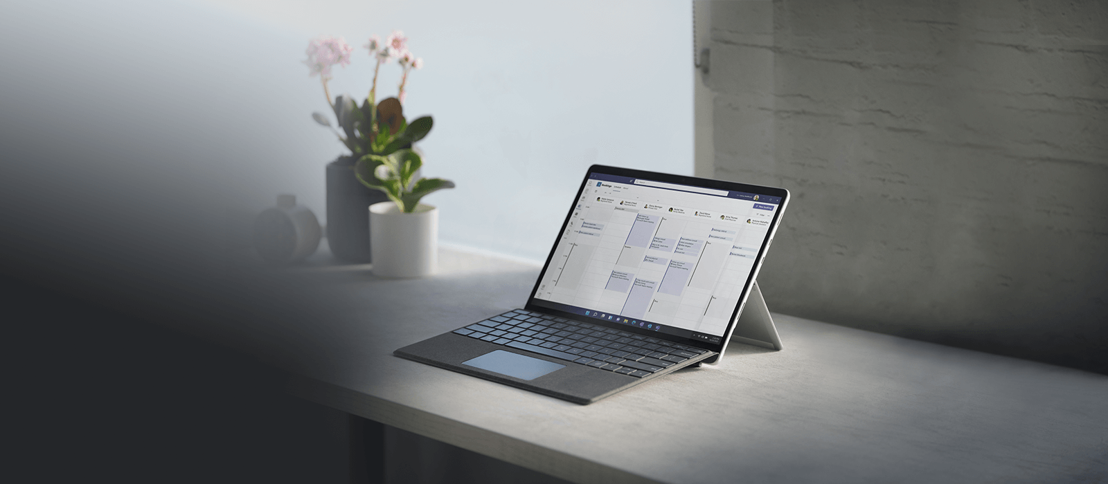 Das Surface Pro 8 steht im Laptop-Modus mit Surface Pro Signature Keyboard auf einem grauen Schreibtisch mit verschiedenen Blumen im Hintergrund
