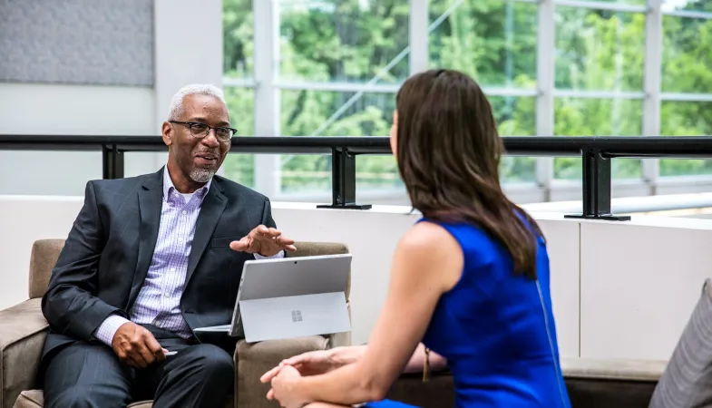 Ein Mann sitzt auf einem Sessel, auf der Lehne hat er das Surface Pro aufgeklappt stehen und erklärt einer Frau gegenüber von sich etwas, die mit dem Rücken zum Betrachter gedreht ist. 