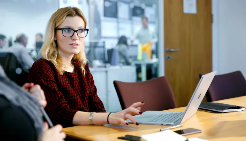 Eine Frau sitzt an einem Tisch in einem Meeting Raum, vor ihr steht das Surface Book aufgeklappt und sie erklärt den Meetingteilnehmern etwas