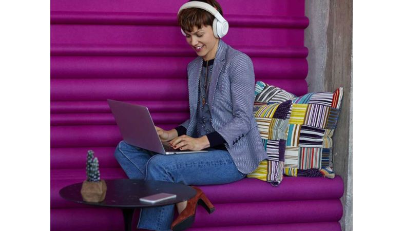 In einer modernen Arbeitsumgebung sitzt eine Frau, die an einem Laptop arbeitet und das Voyager Focus 8200 UC USB-C Headset auf dem Kopf traegt
