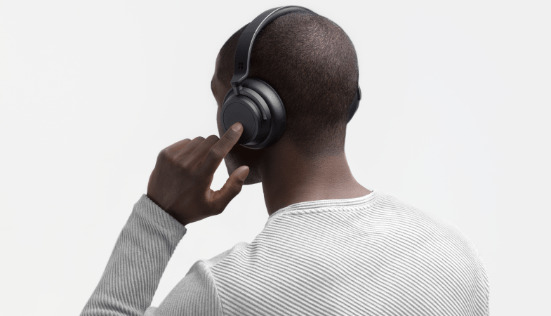 A man wears the Surface Headphones 2 Plus on his head and taps on an on-ear dial 