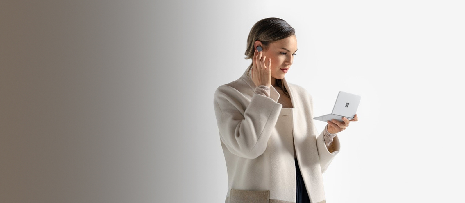 A woman holds the Surface Duo unfolded in one hand, with the other hand she taps on a Surface Earbud she is wearing in her ear
