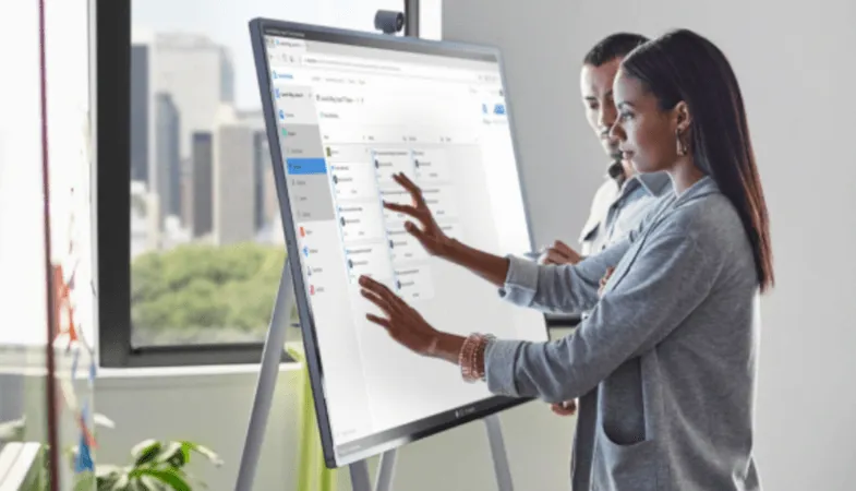 Two people are working on the Surface Hub 2S which is standing on the Steelcase Roam Mobile Stand