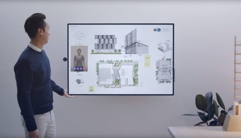 The Surface Hub 2S is hanging horizontally oriented on a wall, a person next to it is looking at the Hub