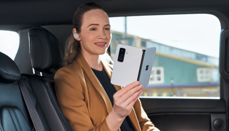 A person sitting in the back seat of a car holding the Surface Duo 2 in the colour Glacier in their right hand