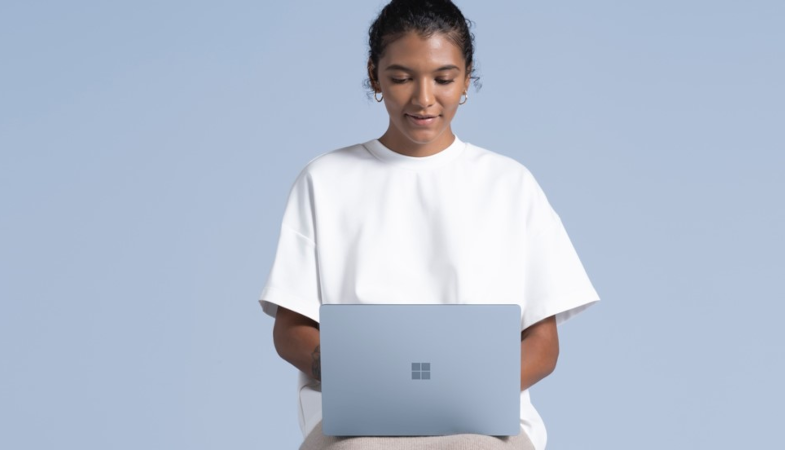 A person is sitting in front of a blue background and has the Surface Laptop 4 in ice blue open on the lap and is typing on the keyboard