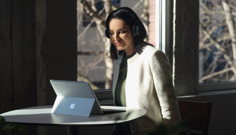 A person sits at a table, wears the Surface Headphones on their head and looks at the screen of the Surface Pro, which is in laptop mode in front of them