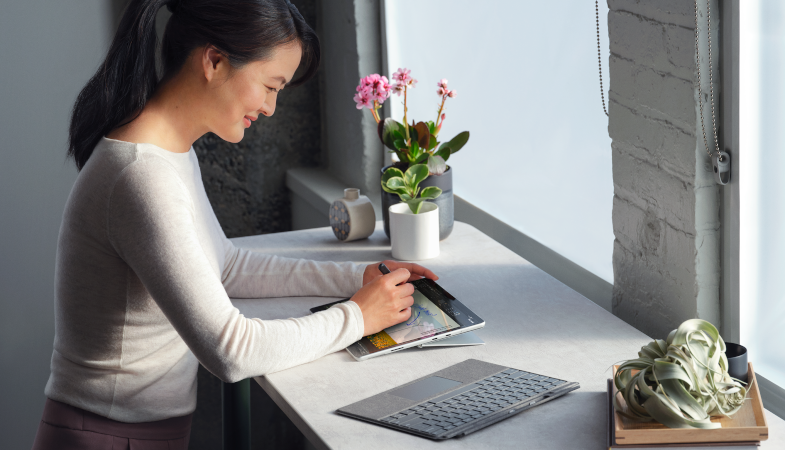 A person writing at a table with the Slim Pen 2 on the Surface Pro 8 display, with the Surface Pro 8 Signature Keyboard next to it