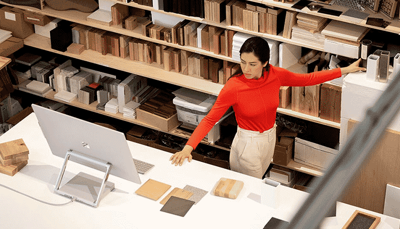 Une femme est debout entre une bibliothèque et un bureau, sur lequel est posé un Surface Studio 2