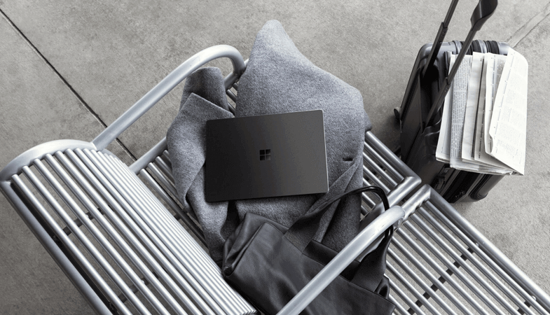 From a bird's eye view, a Surface Laptop lies on a seat in a row of seats in the waiting hall of an airport on a coat. In front of the row of seats is a suitcase with a newspaper on it 