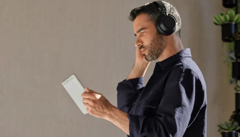 A man holds the Surface Go 3 in tablet mode and uses his right hand to control the Surface Headphones 2+, which he wears on his head. 