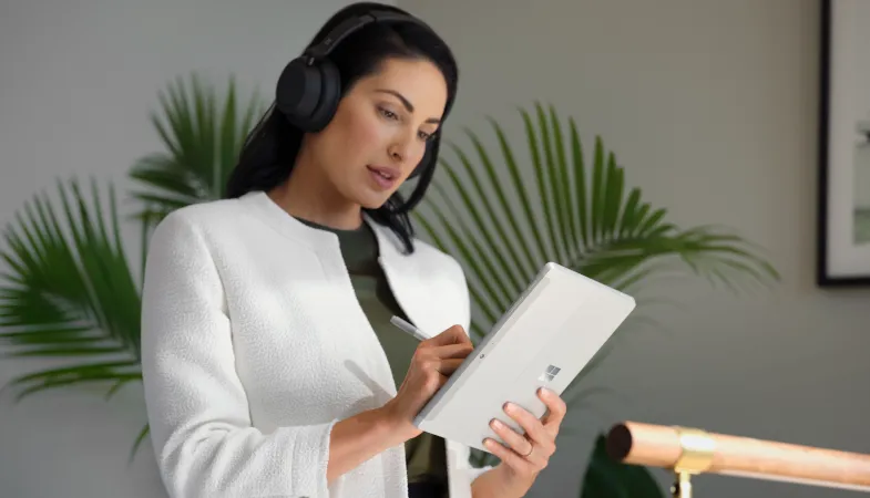 A woman wears the Surface Headphones 2+ on her head and writes with the Surface Pen on the Surface Go 3 she is holding in tablet mode 