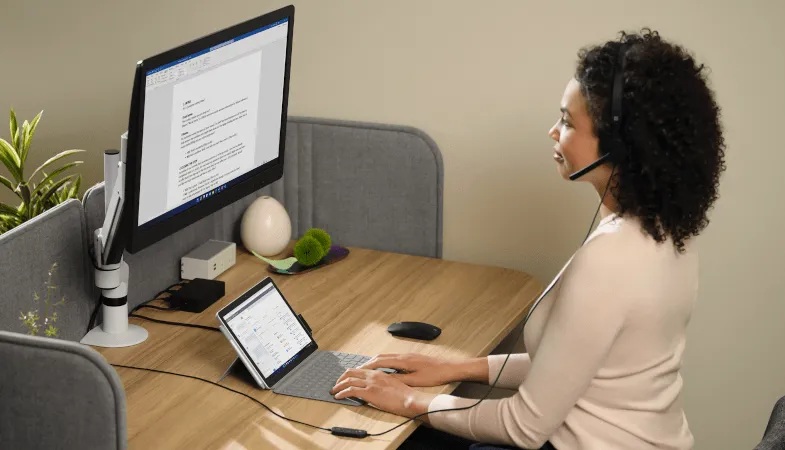 Surface Go 3 for the desktop workplace  A woman sits at a desk and types on the Type Cover of the Surface Go 3 in laptop mode while looking at an external monitor on which a Word document is open