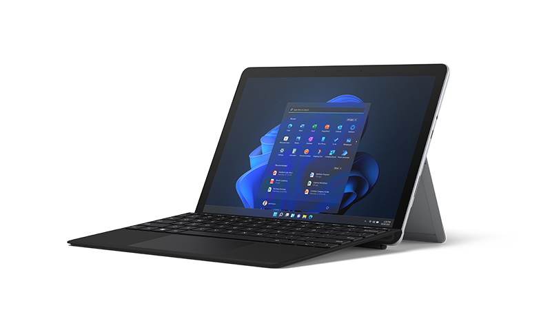 Das Surface Go 3 in Platin inklusive Type Cover in Schwarz  im Laptop-Modus aus seitlicher Perspektive mit blauem Herz links neben dem Surface auf dem NEU steht