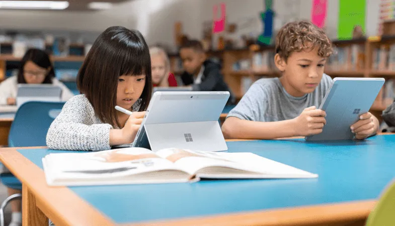 Two children are sitting in a classroom and working on two Surface Go