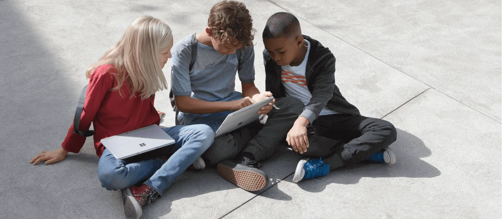 Drei Kinder sitzen auf dem Schulhof und schauen gemeinsam auf den Bildschirm eines Surface Go 