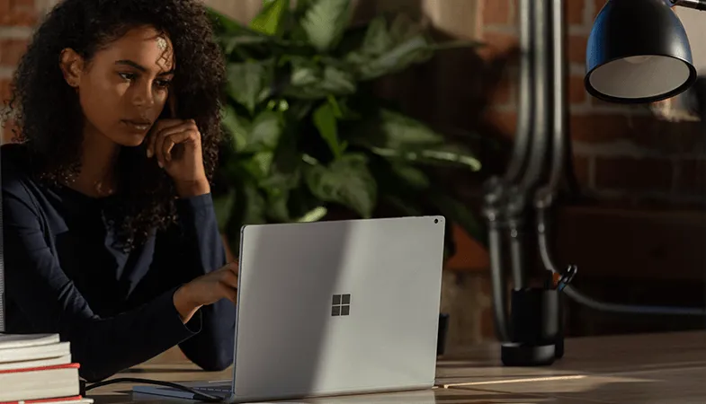 Professionelle Kommunikationsmöglichkeiten über Outlook und Teams in Microsoft 365 Eine Frau sitzt an einem Tisch und arbeitet an einem Surface Book