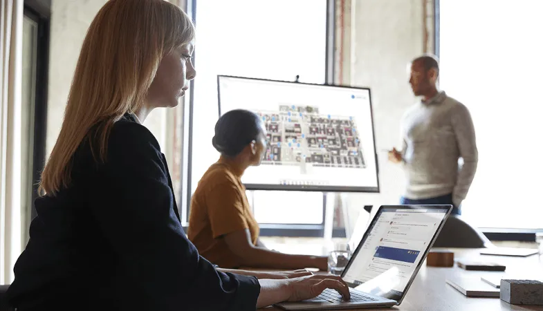 In einem Konferenzraum arbeitet eine Frau am Tisch auf dem Surface Laptop, im Hintergrund zeigt ein Mann einer Frau etwas auf dem Surface Hub 
