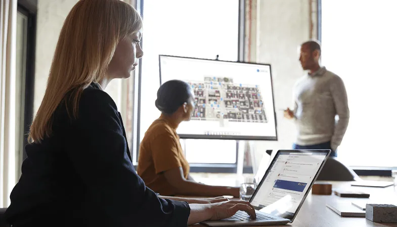Der Arbeitsplatz mit modernster Surface Hardware  In einem Konferenzraum arbeitet eine Frau am Tisch auf dem Surface Laptop, im Hintergrund zeigt ein Mann einer Frau etwas auf dem Surface Hub