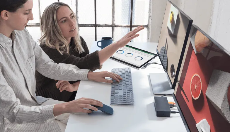 Use Office for collaboration including the benefits of compliance functions  Two women sitting at a desk in front of 2 screens connected to the Surface Pro and working together on a project