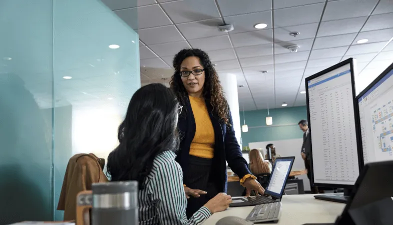 Office 365 E5 als cloudbasierte Komplettlösung für Unternehmen  Eine Frau sitzt am Schreibtisch mit dem Surface Pro und weiteren Bildschirmen auf denen Anwendungen geöffnet sind, eine weitere Frau steht neben dem Schreibtisch und zeigt ihr etwas auf ihrem Surface Laptop