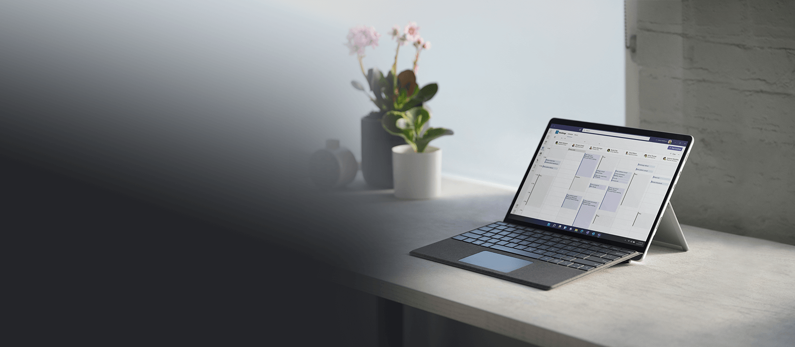 Das Surface Pro 8 steht im Laptop-Modus mit Surface Pro Signature Keyboard auf einem grauen Schreibtisch mit verschiedenen Blumen im Hintergrund