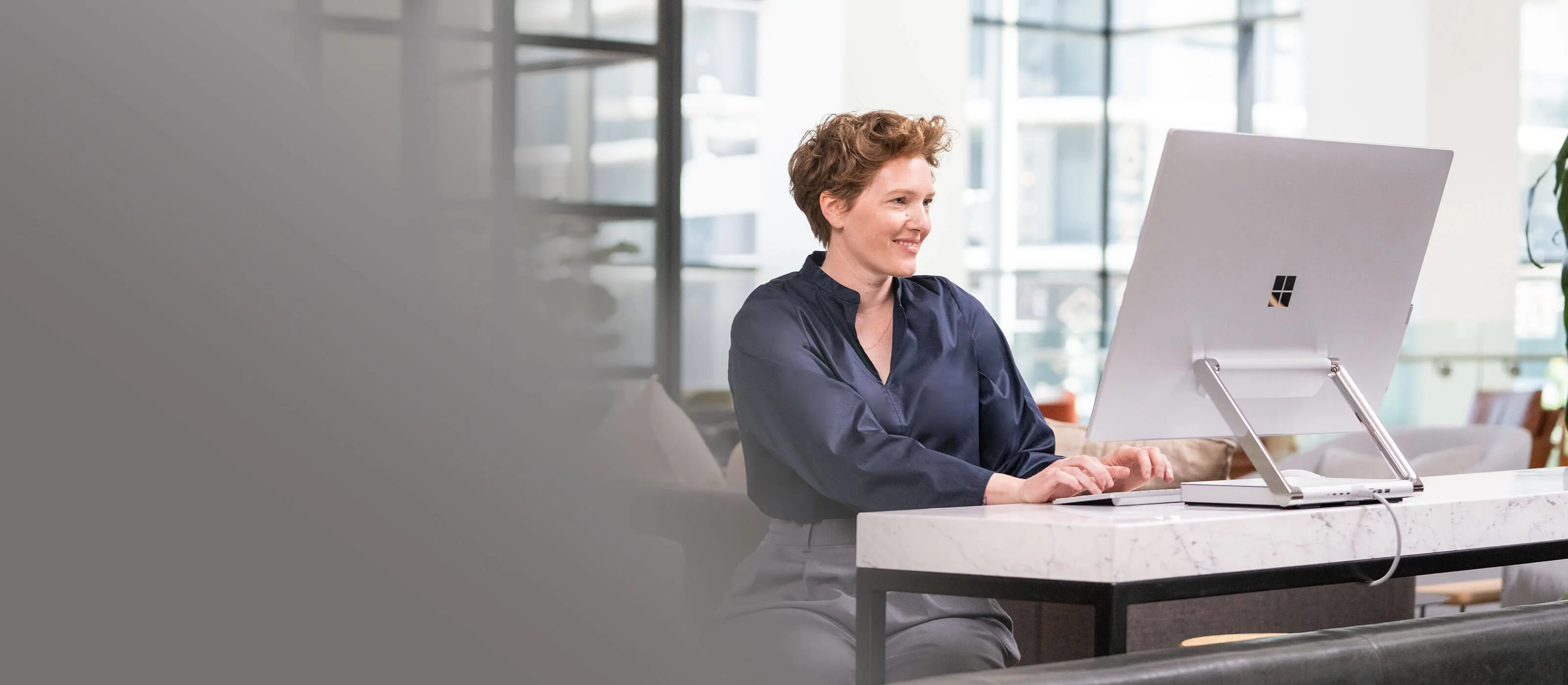 Set your creativity free with the Surface Studio 2+ The Surface Studio 2+ stands on a table, behind it is a woman looking at the Surface Studio and taking something from a shelf