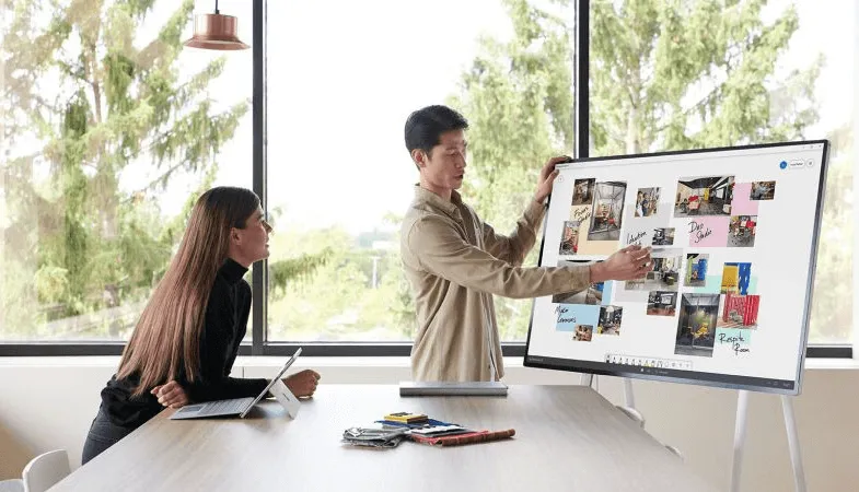 Two people are standing in front of the Surface Hub 2S, one of them is using the Surface Hub 2 Pen 
