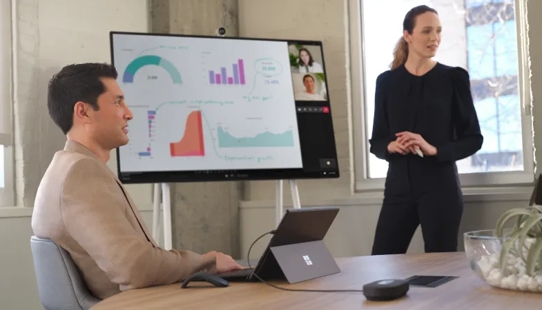 One person is working at a meeting room table on the Surface Pro in laptop mode, while another person is presenting in the background on the Surface Hub.
