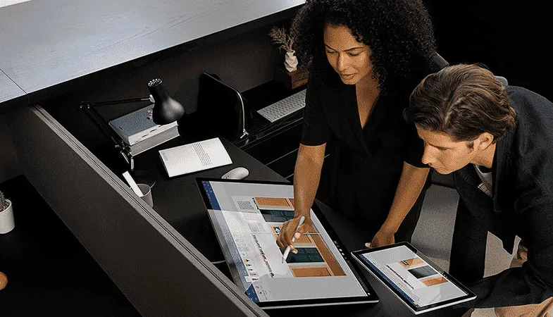 Das Surface Studio 2+ steht in geneigtem Winkel auf einem Scheibtisch, davor stehen zwei Personen und eine von ihnen hält ein Surface Pro