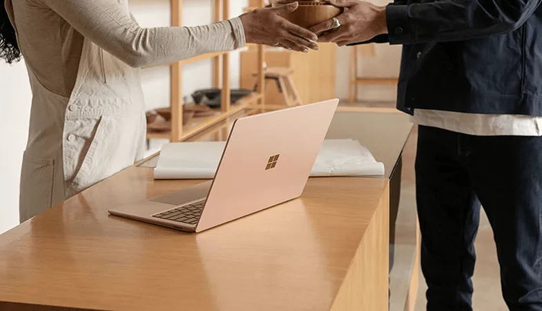 Ein Surface Laptop steht auf einem Tisch, dahinter stehen zwei Personen