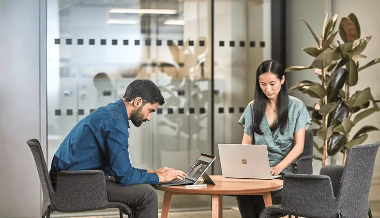 Mit Sway in Microsoft 365 interaktive Webberichte, Präsentationen und vieles mehr gestalten Zwei Kollegen sitzen sich an einem Tisch gegenüber und arbeiten an Surface-Geräten