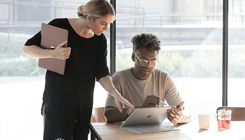 Über die integrierte Teamwebsite SharePoint können Teammitglieder gemeinsam an Projekten arbeiten  Ein Mann sitzt am Tisch und hat ein Surface Book vor sich stehen, neben ihm steht eine Frau, die ihm etwas auf dem Display des Surface Books zeigt