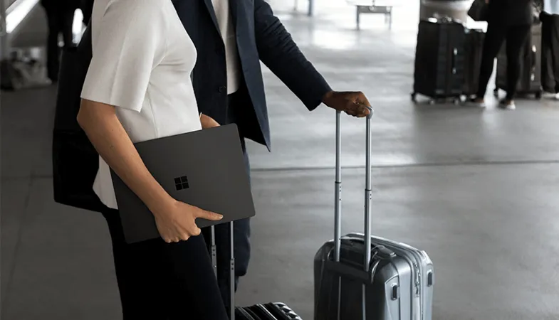 Zwei Personen stehen mit zwei Koffern und einem Laptop in der Hand vor einem Flughafen