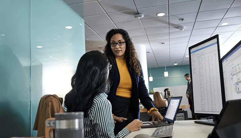 Two women are talking in front of a desk with a Surface Laptop on it