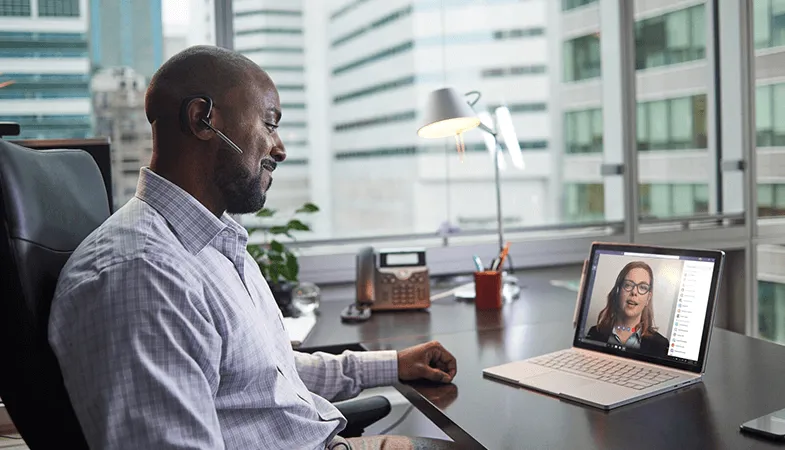 Digitale Zusammenarbeit mit führenden Kommunikationslösungen in Microsoft 365 Ein Mann sitzt am Schreibtisch und führt ein Videotelefonat mit einer Kollegin an seinem Surface Book