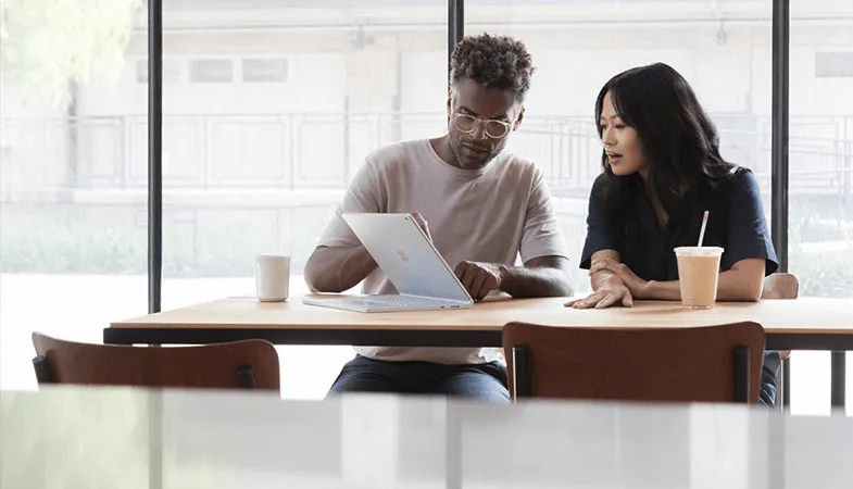 Einfache Windows-Bereitstellung per Windows Virtual Desktop für mehrere Nutzer in Microsoft 365 Zwei Kollegen sitzen nebeneinander am Tisch und schauen auf ein Surface Book