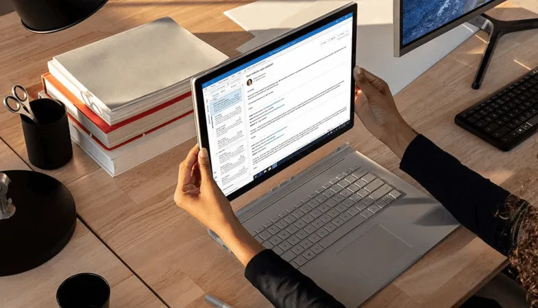 Das Surface Book wird von einer Person an einem Tisch vom Laptop zum Tablet umfunktioniert und das E-Mail-Postfach in Outlook ist geöffnet